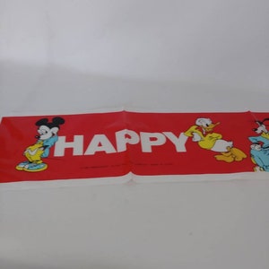May include: A red banner with the word "HAPPY" in white block letters. Mickey Mouse, Donald Duck, and Pluto are featured on the banner. The banner is folded and lying on a white surface.