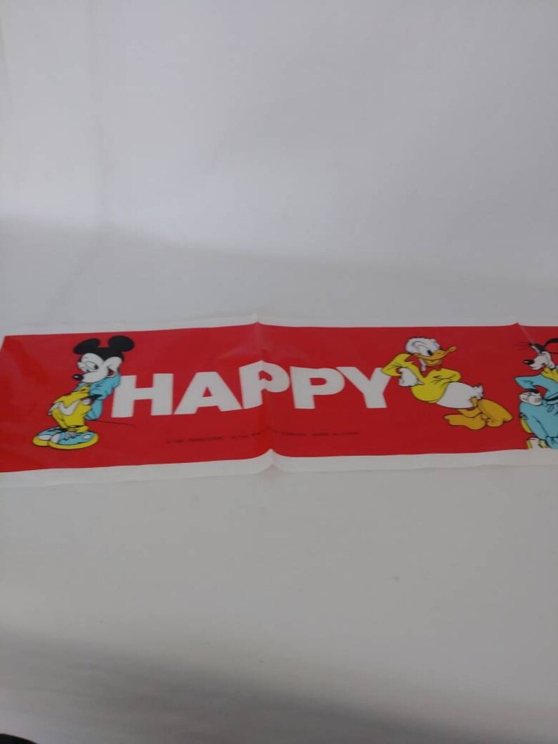 May include: A red banner with the word "HAPPY" in white block letters. Mickey Mouse, Donald Duck, and Pluto are featured on the banner. The banner is folded and lying on a white surface.