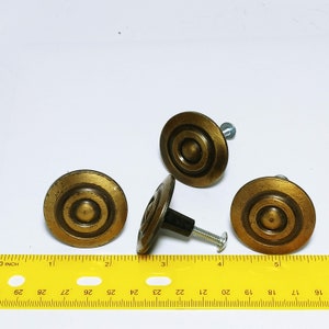 May include: Four antique brass cabinet knobs with a concentric circle design. Each knob has a dark brown stem and a silver screw. A yellow ruler with inch markings is in the foreground.