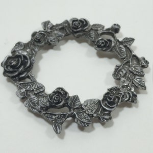 May include: A silver-toned metal wreath featuring detailed rose and leaf designs. The wreath is circular, with roses evenly spaced around the circumference. The leaves and roses have intricate detailing, creating a vintage aesthetic.