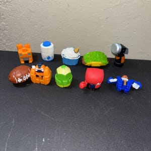 May include: A collection of nine small, colorful plastic toy figures featuring various shapes including a brown football, a green taco, a blue and white milk jug, a red character, a blue and white robot, a green cone, and a silver flashlight.