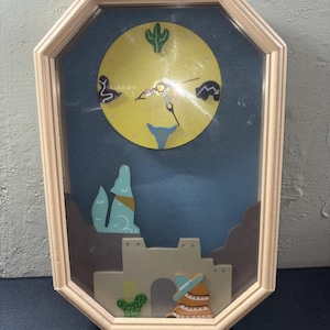 May include: An octagonal wall clock with a light wood frame featuring a yellow circular face with black hands. The background depicts a desert scene with a blue sky, brown mountains, a light blue howling coyote, a tan adobe structure, and cacti.