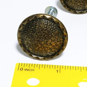 May include: Two antique brass cabinet knobs are shown. Each knob has a textured, circular face with a decorative edge. The knobs are approximately 2.5 cm in diameter. A yellow ruler is in the foreground.