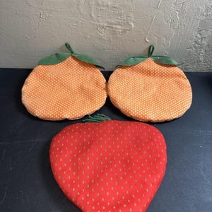 May include: Three fruit-shaped pot holders. Two are orange with white polka dots and green stems. The third is red with gold dots, resembling a strawberry. These kitchen accessories add a touch of whimsy to any cooking space.