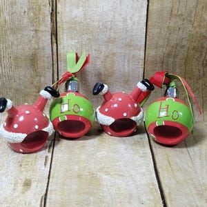 May include: Three red and green Christmas baubles shaped like Santa Claus with his arms and legs in the air. The baubles have white polka dots and a white fur trim. Each bauble has the word "Ho" written on it.