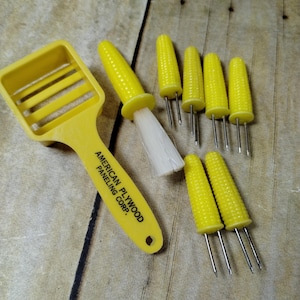 May include: Yellow corn on the cob holders with a yellow corn cob shaped brush and a yellow plastic corn cob cutter. The corn cob holders have three prongs and the text "AMERICAN PLYWOOD PANELING CORP." is printed on the cutter.