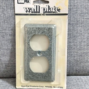 Puede incluir: Una placa de pared de metal plateado con una sola abertura de tomacorriente. El empaque dice "Gam-Pak wall plate" y "Gam-Pak Products Corp., Hillside, N.J. 07205".
