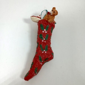 May include: A red Christmas stocking ornament with green holly and white berry accents. A small brown teddy bear and angel wings peek out from the top. The ornament is designed for holiday decor.