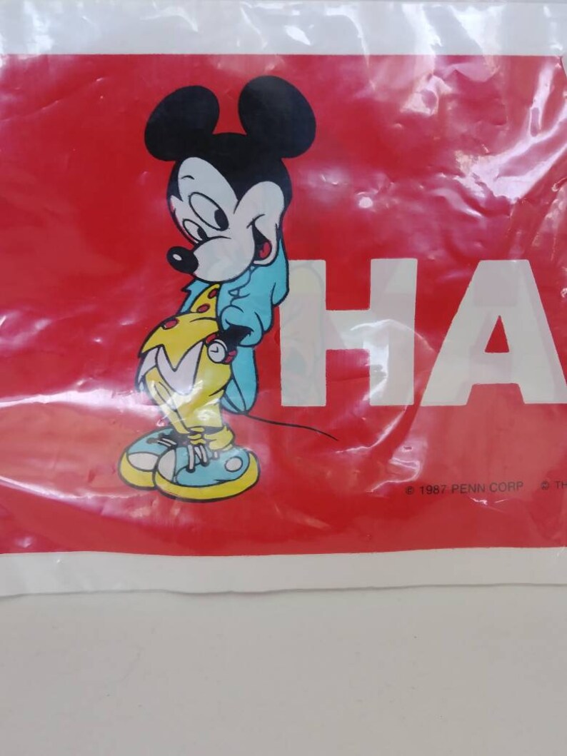 May include: A red plastic bag featuring a cartoon image of Mickey Mouse in a blue jacket and yellow shoes. The word "HA" is printed in white. The bag has a copyright notice from 1987.