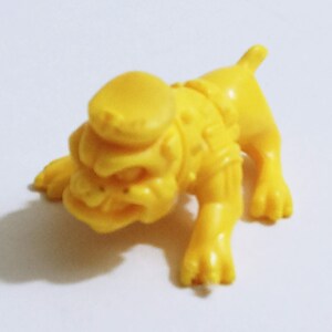 May include: A small, yellow plastic toy dog in a crawling position. The dog has a large head with a hat, a textured body, and four legs. The toy is made of a solid, bright yellow plastic.