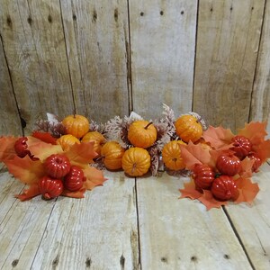 May include: A fall-themed arrangement featuring miniature orange pumpkins and red gourds. The pumpkins are nestled among orange maple leaves and dried floral accents, set against a rustic wooden background.
