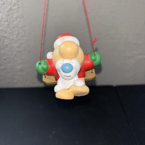 May include: A hanging ornament featuring a cartoon character with a large tan nose, a blue nose tip, and a white beard, wearing a red jacket with green sleeves and a red hat. The character sits on a small tan swing suspended by thin red strings.