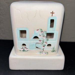 May include: A cream-colored ceramic luminary shaped like a mission-style building. The structure features light blue painted window frames, a brown cross on the tower, and hand-painted floral vines climbing the front facade with small green plants.