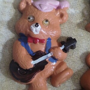 May include: A brown teddy bear wearing a pink hat and a blue shirt plays a brown guitar with white strings. The bear has white eyes and a white nose.