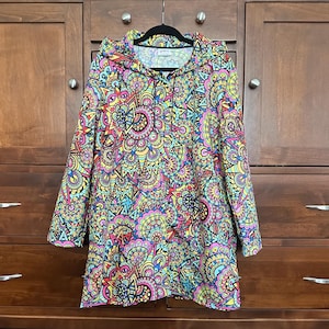 May include: A long-sleeved hoodie with a vibrant, intricate pattern in shades of pink, yellow, blue, and green. The design features a repeating, mandala-like motif. The hoodie is hanging on a black hanger against a wooden cabinet.