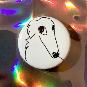 May include: A white button with a black line drawing of a dog's head. The dog has long features and a small black nose. The button is round and has a glossy finish. The background is a holographic, iridescent surface.