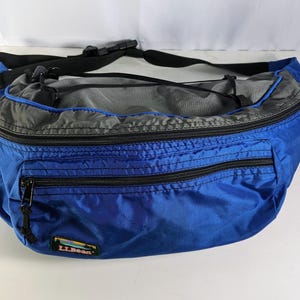 May include: A blue and gray fanny pack with black zippers and a black adjustable strap. The bag has a rectangular shape with multiple zippered compartments. The L.L.Bean logo is visible on the front.
