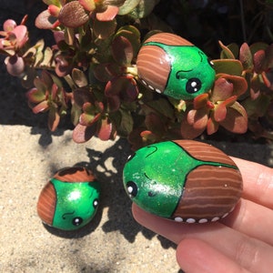 May include: Hand-painted decorative rocks shaped like cartoon beetles. Each rock features a green head and a brown body with white accents. The rocks are displayed near a succulent plant, creating a whimsical, nature-inspired scene.