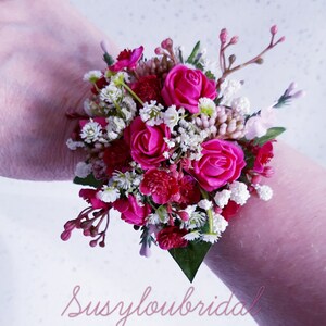 May include: A pink and white floral wrist corsage with pink roses and white baby's breath.  The corsage is tied with a white ribbon and is perfect for a wedding or special occasion.