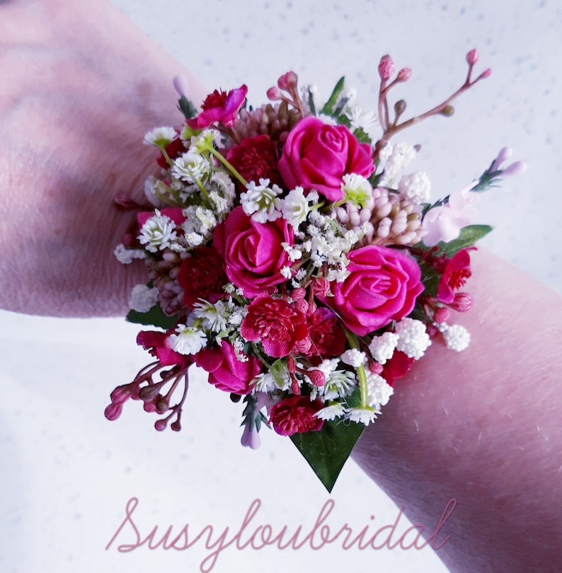 May include: A pink and white floral wrist corsage with pink roses and white baby's breath.  The corsage is tied with a white ribbon and is perfect for a wedding or special occasion.