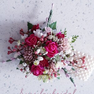 May include: A small bouquet of pink and white flowers with pearl accents. The bouquet is arranged in a circular shape and features a variety of flowers, including roses, baby's breath, and other small flowers. The bouquet is accented with a string of pearls.
