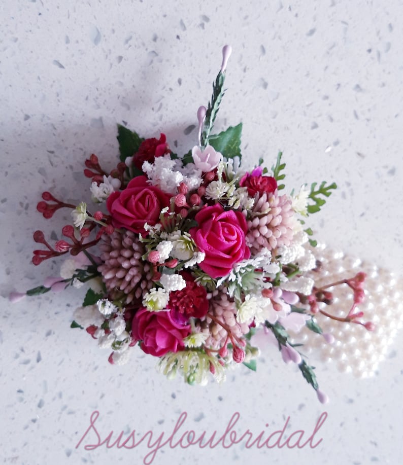May include: A small bouquet of pink and white flowers with pearl accents. The bouquet is arranged in a circular shape and features a variety of flowers, including roses, baby's breath, and other small flowers. The bouquet is accented with a string of pearls.