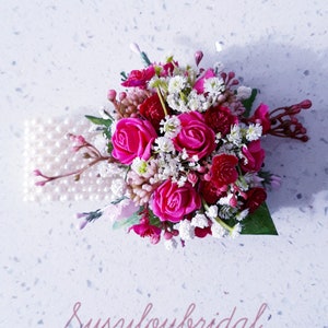 May include: A pink and white floral bridal wrist corsage with pearl accents. The corsage features pink roses and white baby's breath.