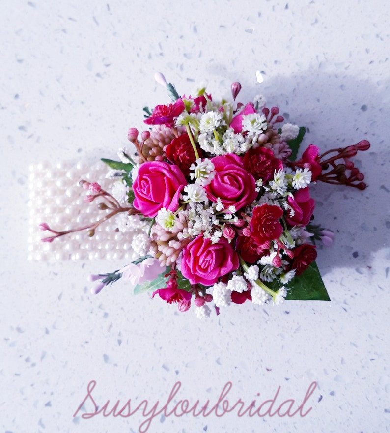 May include: A pink and white floral bridal wrist corsage with pearl accents. The corsage features pink roses and white baby's breath.