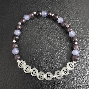 Può includere: Un braccialetto di perline con la parola "ELDERMO" scritta con perline di lettere bianche. Il braccialetto presenta perline viola scuro e blu chiaro alternate, con piccole perline distanziatrici argentate. Il braccialetto è su una superficie nera.