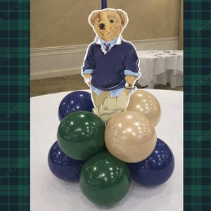 Könnte beinhalten: Dekorative Tischdekoration mit einem Bären-Ausschnitt in einem marineblauen Pullover und khakifarbener Hose, auf einer Gruppe von marineblauen, grünen und beigefarbenen Luftballons. Der Hintergrund umfasst eine weiße Tischdecke und ein Karomuster.