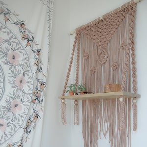 May include: A white pillow with black text that reads "VIRGO" and a small pink zodiac symbol. The pillow is on a bed with a pink blanket and a white wall behind it. A wooden shelf with a macrame wall hanging is on the wall.