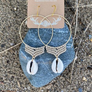 May include: A pair of gold hoop earrings with macrame and shell accents. The earrings are handmade and feature a natural shell charm hanging from each hoop. The earrings are displayed on a brown card with the text "BESPOKE MACRAME bespokemacrame.etsy.com"
