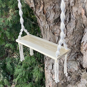 Op de afbeelding: Een houten plank met een natuurlijke afwerking hangt aan een tak van een boom met twee witte macramé touwen met kwastjes. De plank is ongeveer 40 cm lang en 10 cm breed.