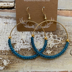 Puede incluir: Pendientes de aro de macramé de color turquesa con aros dorados y cuentas doradas. Los pendientes se muestran en una tarjeta marrón con el texto "BESPOKE MACRAME bespokemacrame.etsy.com"