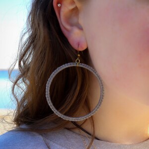 May include: A pair of large hoop earrings made of gray woven cord. The earrings are hanging from the ear of a person wearing a gray sweater.