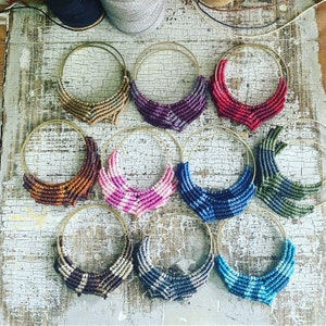 May include: A collection of handmade macrame hoop earrings in various colors, including brown, purple, red, orange, pink, blue, and green. The earrings feature gold-colored hoops and intricate woven designs, showcasing a bohemian style.