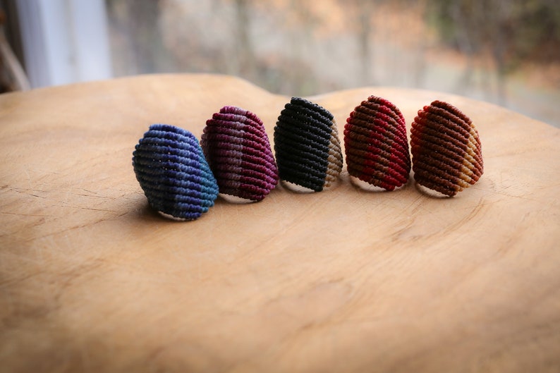 K&ouml;nnte beinhalten: Sechs bunte gewebte Ringe, jeder mit einer anderen Farbkombination. Die Ringe sind aus einem dicken, strukturierten Material gefertigt und in einer Reihe auf einer Holzoberfl&auml;che angeordnet.