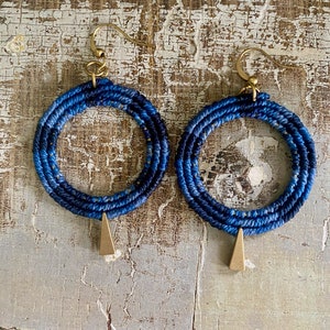 May include: A pair of blue and gold hoop earrings with a triangle pendant. The hoops are made of woven fabric and the pendant is made of metal.