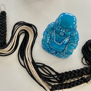 May include: A black and cream macrame plant hanger with two metal rings at the top. The hanger is hanging over a blue ceramic statue of a smiling Buddha.