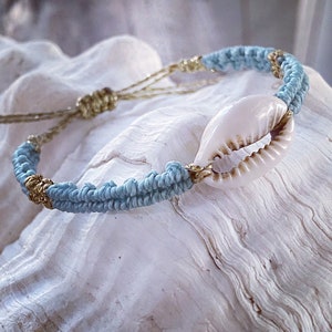 Pode incluir: Uma pulseira trançada azul claro e dourada com um amuleto de concha de cauri branco.