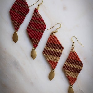 May include: Four pairs of handmade diamond-shaped earrings with a woven pattern in shades of red, brown, and beige. Each earring features a gold-toned leaf-shaped dangle.
