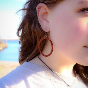 May include: A close-up of a person's ear wearing a pair of red hoop earrings. The earrings are made of woven material and have a brass hook.
