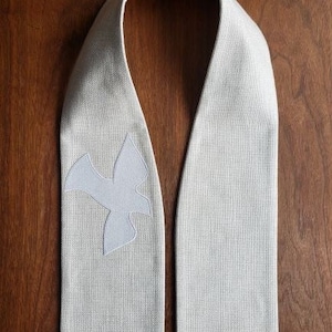 May include: A white linen stole with a white dove embroidered on one side. The stole is a long, narrow piece of fabric worn by clergy during religious services.