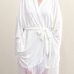 May include: White robe with a long sleeve and a fringe trim. The robe has a sash tie at the waist.