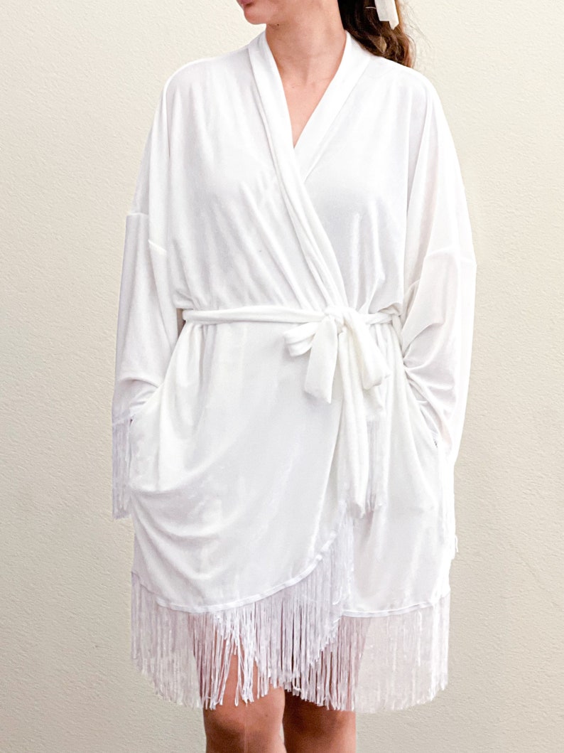 May include: White robe with a long sleeve and a fringe trim. The robe has a sash tie at the waist.