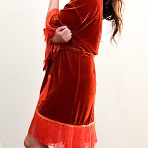May include: A rust-colored velvet robe with a fringe trim. The robe has a tie belt and long sleeves.