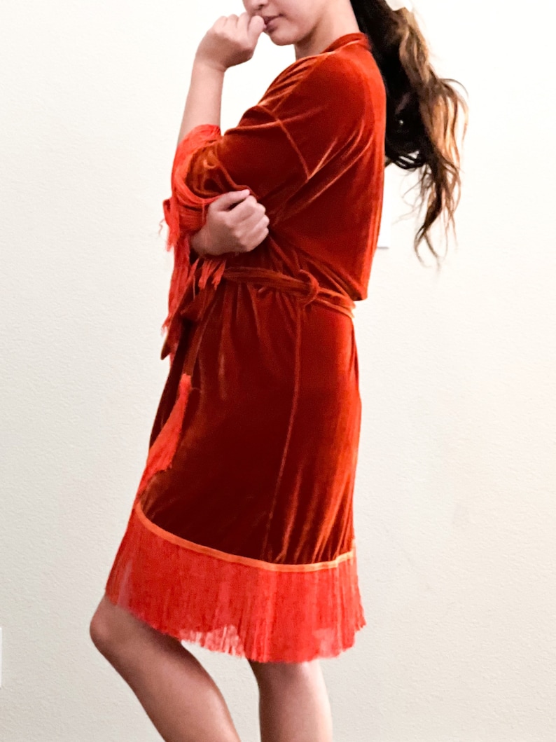 May include: A rust-colored velvet robe with a fringe trim. The robe has a tie belt and long sleeves.