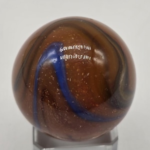 Könnte beinhalten: Eine braune Murmel mit blauen und schwarzen Wirbeln. Die Murmel hat eine glänzende Oberfläche und kleine weiße Flecken. Die Murmel liegt auf einem transparenten Ständer.