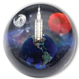 May include: A glass paperweight with a silver rocket ship launching from a blue and green Earth. The background is a dark blue space with a white moon and a red Mars.