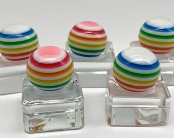 May include: Five rainbow striped glass marbles on clear square bases. Each marble has a white base and is decorated with alternating stripes of red, orange, yellow, green, blue, and pink.
